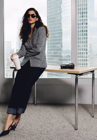 Blazer a motivo houndstooth bianco e nero, jeans neri con orli arrotolati, borsa nera, tazza di caffè bianca, in un ambiente d'ufficio con grandi finestre.