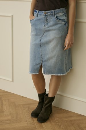 Žena stojící opřená o krémově zbarvenou zeď, má na sobě světle modrou džínovou sukni ke kolenům s předním rozparkem a černé kotníkové boty.