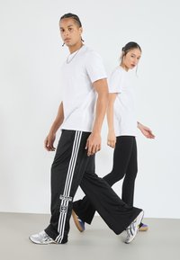 Juoda sportinė kelnės su baltais dryžiais ir logotipu; balta marškinė su apvalia iškirpte. Modelis vilki juodas leginsus ir mišrių spalvų sportinius batus.