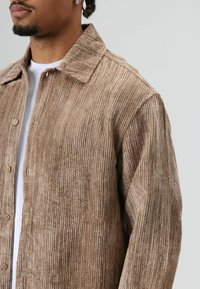 Brown corduroy shirt with a textured, ribbed design, featuring a pointed collar and button closure. Worn over a white shirt.