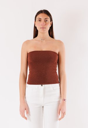 Giovane donna con lunghi capelli scuri, che indossa un top marrone senza spalline e pantaloni bianchi, in piedi davanti a uno sfondo bianco neutro.