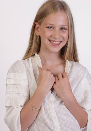 Gouden sieraden met een hartvormige hanger, armband met een hartcharme, gedragen met een gestructureerde witte blouse en subtiele ruches.