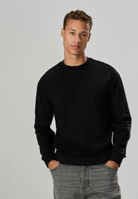 Sort crewneck sweatshirt med lange ærmer, lavet af et blødt materiale. Modellen har hænderne i lommerne og har den parret med grå jeans.