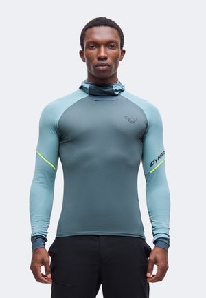 Homme portant un haut de sport ajusté bleu sarcelle à manches longues avec col montant et pantalon noir, debout devant un fond clair uni.