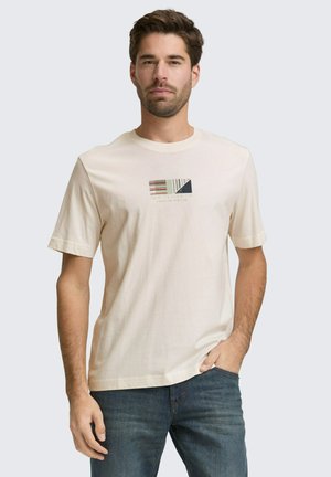 Mann trägt cremefarbenes kurzärmeliges T-Shirt mit mehrfarbigem geometrischem Muster und blaue Jeans, steht mit einer Hand in der Tasche vor einfarbigem Hintergrund.