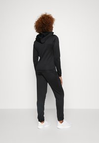 Čierna mikina s kapucňou a súprava na jogu v hladkej, elastickej tkanine. Obsahuje podšívku kapucne a elastický pás a manžety. Nosené s bielymi športovými topánkami.