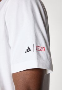 Hvit bomull t-skjorte med korte ermer, et svart Adidas-logo og et rødt Marvel-merke på venstre erme. Glatt tekstur.