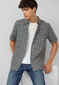 Junger Mann in blau kariertem Kurzarmhemd über weißem T-Shirt und Jeans, sitzt auf weißem Klotz, schaut nach links vor schlichtem grauem Hintergrund.