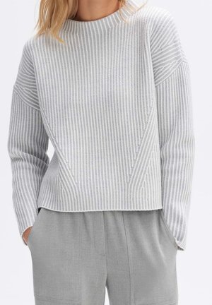 Kobieta ubrana w jasnoszary prążkowany sweter i pasujące jasnoszare spodnie z rękami w kieszeniach, zdjęcie kadrowane od szyi do połowy ud.