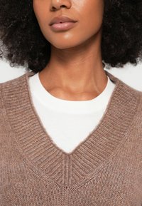 Camisola castanha de malha com decote em V profundo sobre uma t-shirt branca. A textura é suave com detalhes canelados no pescoço.