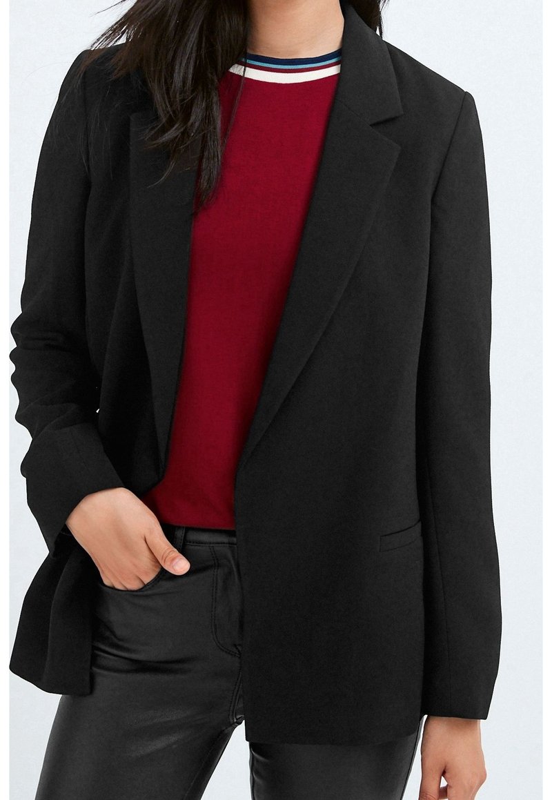 Blazer noir à coupe ajustée, avec des revers en cranté et deux poches avant. Porté sur une chemise rouge avec un col rayé. Texture lisse.