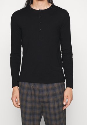 Personne portant une chemise henley noire ajustée à manches longues avec trois boutons et un pantalon ample à carreaux dans des tons bleus et bruns.