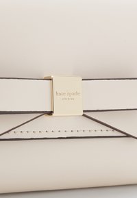 White leather wallet with dark brown accents and stitching. Features a gold clasp with "kate spade" engraving. Flat rectangular design.