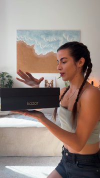 Fekete téglalap alakú doboz arany "kazar" logóval, amit egy személy tart. A doboz sima, matt felülettel rendelkezik. Természetes fény világítja meg a jelenetet.