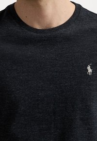 Man som bär en mörkgrå melerad t-shirt med rund halsringning och en liten vit polo-spelare broderad på vänster bröst.