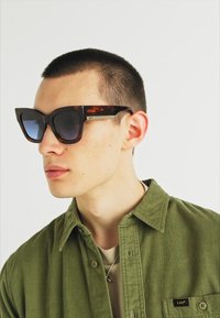 Schildpad patroon cat-eye zonnebril met donkere lenzen en metalen accenten, gedragen door een persoon in een olijfgroen shirt tegen een witte achtergrond.