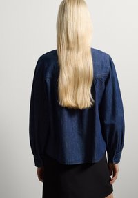 Donkerblauwe denimshirt met lange mouwen, een relaxte pasvorm en knoopmanchetten, gedragen met een zwarte rok, gezien van achteren. Lange, steile haren.