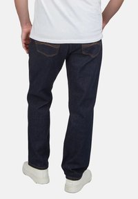 Jeans de mezclilla azul oscuro con corte recto, costuras visibles y dos bolsillos traseros. Llevados con zapatillas blancas y una camiseta blanca lisa.