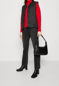 Gilet matelassé noir par-dessus un pull à col roulé rouge, associé à un jean noir droit et des bottines à talons block. Accessoirisé avec un sac à main noir texturé.