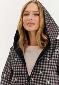 Jauna sieviete, tērpta melnā un rozā šaha raksta kapuci jakā ar krāsainiem sirds rakstiem virs bēša augšpusē, nedaudz smaidot.