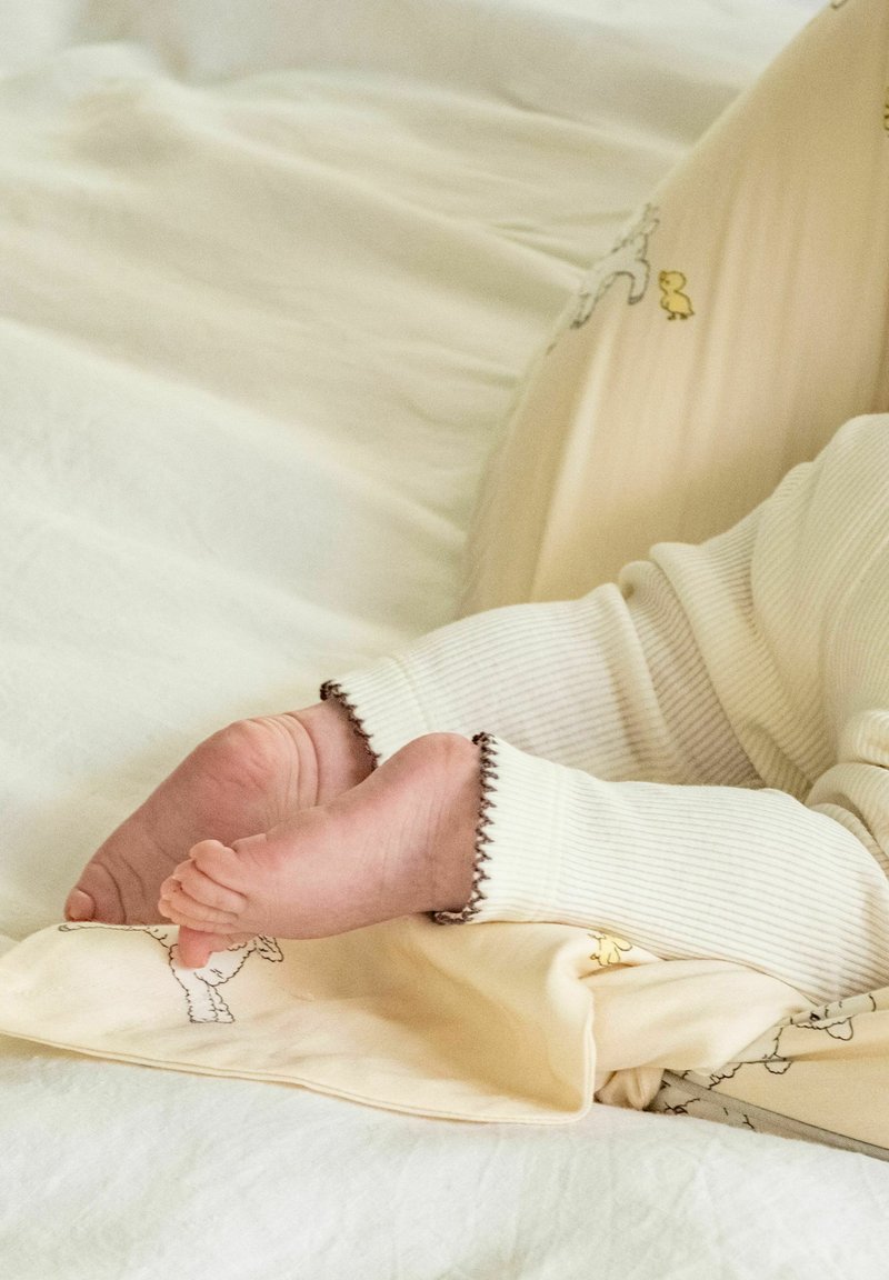 Pieds de bébé en pyjama côtelé couleur crème avec bordure foncée, reposant sur une couverture jaune clair avec des motifs d’animaux sur un matelas blanc.