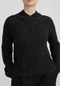 Black, textured sweater with a high neckline and long sleeves. Fuzzy material with a loose fit and a subtle pattern, layered over black trousers.