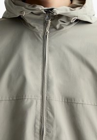 Veste légère grise, résistante à l'eau, avec un zip à l'avant, une capuche réglable et une texture lisse. Présente des lignes de couture et des accents à cordon.