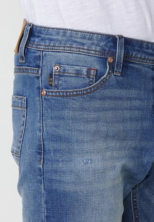 I jeans in denim blu presentano una vestibilità dritta, un lavaggio chiaro, una leggera sfumatura, e finiture in rame con diverse tasche e dettagli del logo.