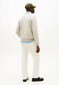 Mand iført en beige sweatshirt, lyseblå skjorte, hvide bukser, sorte sko og en olivengrøn kasket, stående sidelæns mod en hvid baggrund.