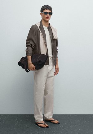 Brown zip-up jacket with a beige collar, cream t-shirt, light beige trousers, black sunglasses, and a dark tote bag. Black flip-flops.