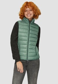 Grüner gepolsterter Weste mit hohem Kragen, Reißverschluss und Seitentaschen. Kombiniert mit einem schwarzen strukturierten Langarmoberteil. Graue Jeans.