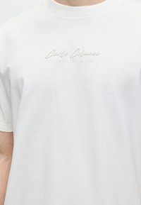 Λευκό κοντομάνικο μπλουζάκι με μικρό ανάγλυφο κείμενο στο στήθος που αναγράφει "Carlo Colucci A Legacy Since 1978".