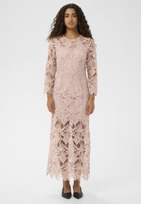 Rochie din dantelă de culoare roz prăfuit, cu mâneci lungi și motive florale, cu un design până la podea și talie ajustată. Purtați cu pantofi cu vârful ascuțit, negri.