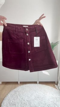Bordo kordna krilo s sprednjimi gumbi in dvema velikima žepoma, ki ga držijo roke, cenjeno z priloženo belo oznako.
