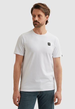 Man met baard die een eenvoudig wit T-shirt met korte mouwen draagt met een klein zwart logopatch op de borst, staande tegen een lichtgrijze achtergrond.