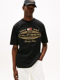 Svart T-skjorte med korte ermer som har et gull grafisk design med teksten "HILFIGER Racing Team" og en logo. Brukt med lyse bukser og en matchende caps.