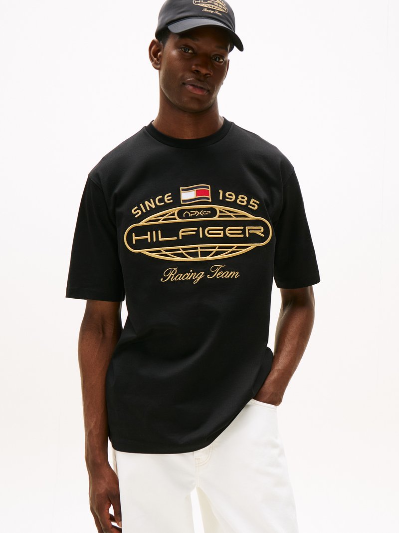 Svart T-skjorte med korte ermer som har et gull grafisk design med teksten "HILFIGER Racing Team" og en logo. Brukt med lyse bukser og en matchende caps.