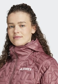 Padded, wysokiej szyi kurtka w kolorze mauve z pikowaną fakturą. Posiada zamek błyskawiczny z przodu oraz biały logo "TERREX" na klatce piersiowej.