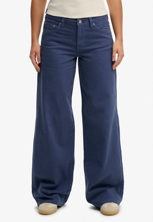 Osoba nosząca granatowe spodnie o wysokim stanie, szerokich nogawkach z pięcioma kieszeniami oraz beżowe trampki na białym tle.