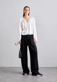 Weiße Bluse mit langen Ärmeln und V-Ausschnitt sowie gerafftem Schulterdetail, kombiniert mit lockeren schwarzen Hosen und flachen, zweifarbigen Schuhen. Schwarze Handtasche.