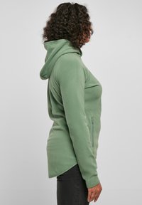 Green fleece hoodie featuring a zippered pocket, long sleeves, and a rounded hem. Soft texture and fitted silhouette.