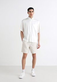 Chemise blanche à manches courtes avec une poche, associée à un short beige clair. Stylée avec des baskets blanches, sur un fond uni.