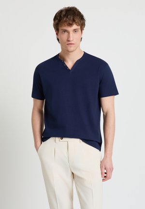 Giovane uomo con indosso una maglietta henley blu navy a maniche corte e pantaloni plissettati color crema, con le mani nelle tasche, in piedi davanti a uno sfondo chiaro e uniforme.