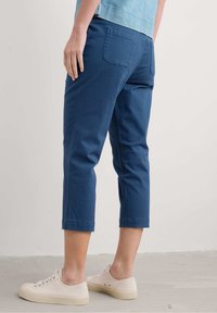 Könnyű pamutból készült kék cropped nadrág, sima textúrával, két hátsó zsebbel és felgöngyölített szegélyekkel. Bézs sneaker cipővel párosítva.