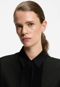 Camicia nera con bottoni, colletto strutturato e texture plissettata sul davanti. Tessuto liscio con finitura lucida.