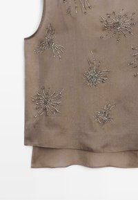 Mouwloze beige top met gelaagde zoom, versierd met zilverkleurige bloem- en stervormige kralenversieringen verspreid over de stof.