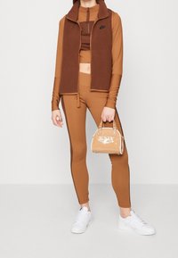 Hnědá fleecová vesta přes odpovídající dlouhý rukáv a legíny, doplněná bílými teniskami a malou krémovou kabelkou se značením.