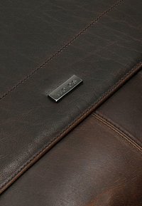 Hnědá kožená peněženka s texturovaným povrchem, zdobená stříbrnou kovovou logo destičkou a kontrastním prošíváním podél okrajů.
