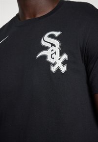 Camiseta de algodón negra con un logotipo blanco prominente y un diseño curvado, con letras distintivas en el área del pecho izquierdo.