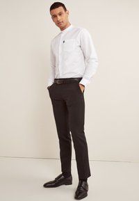 Hvid skjorte med knapper og mandarin-krave samt brystlomme på venstre side, kombineret med sorte, tilspidsede bukser og sorte pæne sko.